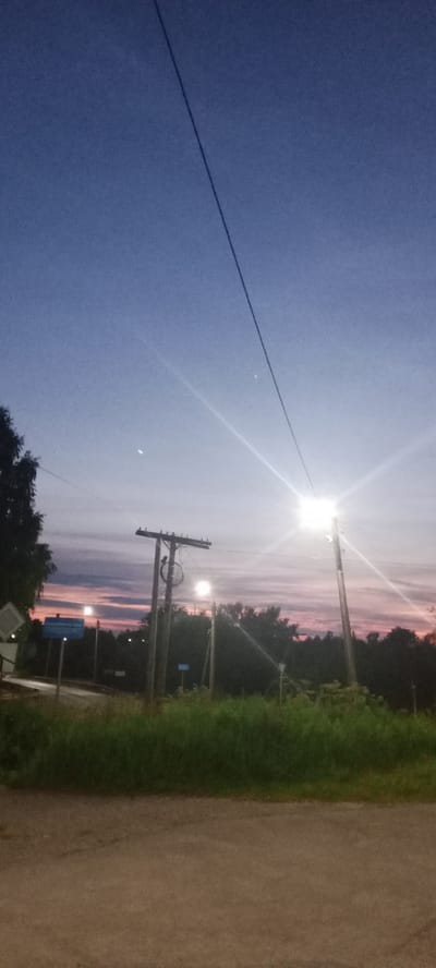This media file depicts a rural roadside scene at dusk in Kudymkar, Russia. The sky transitions from a deep blue at the top to lighter shades of purple and pink near the horizon, indicating the twilight hours. A single power line stretches diagonally across the frame from the upper left to the lower right.
Prominently featured are two utility poles with insulators. The pole on the left has a circular object, possibly a tire, hanging from it. Both poles are illuminated by bright, artificial lights, suggesting the scene is near a road or settlement. In the background, lush green grass and trees form a dense line, with some darker shapes suggesting buildings or structures amidst the foliage. A road is visible in the foreground, with a signpost on the left side of the frame.
There are no people visible in the image. The overall mood is quiet and serene, characteristic of a rural setting at the end of the day. The presence of streetlights and the road suggests this is likely a point along a rural highway or a road within a small community. The weather appears clear, with the colors in the sky suggesting a calm evening. A small, bright object, possibly a planet or bright star, is visible in the upper portion of the sky, along with a fainter, crescent-shaped object.