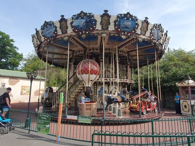 The image shows a carousel ride in a park, likely a fairground or amusement park. The carousel is decorated with ornate details and features various animals and vehicles for children to ride. In the foreground, a green metal fence encloses the ride. To the left of the carousel, a man wearing a black t-shirt and shorts is standing near a baby stroller, looking towards the carousel. A sign on the fence reads "Le Carrousel de Jules," indicating the name of the ride. Behind the fence, trees and foliage suggest an outdoor environment. The carousel itself has a large hot air balloon in the center, surrounded by poles and decorative elements. Several children are visible on the carousel, some riding on horses and others in a red fire truck. The sky is blue with some clouds, indicating a daytime scene. A building with a light-colored brick facade and a green roof is visible in the background. A lamppost stands near the entrance of the carousel area. The overall impression is of a cheerful, family-friendly attraction. The sign "Le Carrousel de Jules" suggests the carousel is located in or is associated with Bretteville-sur-Odon, France.