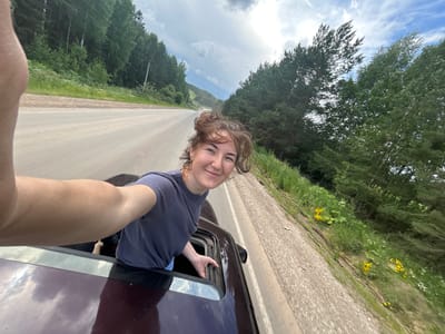 A young woman with messy brown hair, wearing a dark blue t-shirt and denim shorts, leans out of a car sunroof while smiling and holding the frame. Her left arm is extended, holding the camera for a selfie. The car appears to be on a paved road with a gravel shoulder, bordered by a dense forest of green trees and some yellow wildflowers. The sky is partially cloudy with patches of blue. The image is taken from a high angle, looking down and to the right. The context of the city of Верх-Рождество, Russia is inferred from the provided information, though not explicitly visible in the image itself.