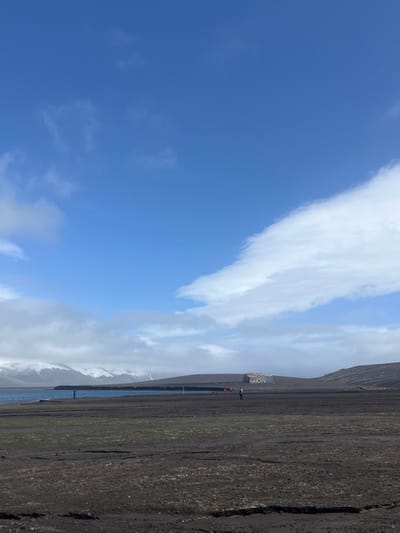 A landscape photograph captures a bright blue sky with scattered white clouds over a dark, volcanic terrain. A strip of water separates the land from a distant, snow-capped mountain range obscured by cloud cover. A building is visible atop the dark ground, with small figures and orange markers positioned near the water's edge.