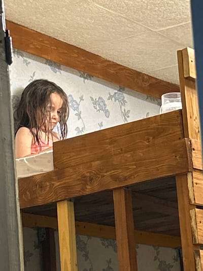 A young girl with wet, dark hair lies down on what appears to be a bed. Her head is propped up slightly, and her eyes are closed or downcast. She is wearing a pink tank top. The background consists of light-colored, patterned wallpaper with a floral design in shades of blue and gray. Above the girl, there are wooden beams and planks, suggesting an attic or a structure with a sloped ceiling. A clear plastic container is visible to the upper right of the girl. The lighting is dim, and the overall mood appears calm and possibly sleepy. The image seems to have been taken indoors.