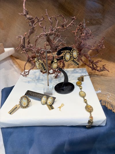 This image shows a display of ornate jewelry, likely in a boutique or market stall in Würzburg, Germany. The foreground features a white rectangular platform covered with a blue cloth draped over its left side, serving as a display surface. On this platform, several pieces of jewelry are arranged.

To the left, a ring with an oval, patterned centerpiece surrounded by small amber-colored stones is displayed with a price tag reading "KONPLOTT 54,90 €". Next to it, a pair of matching oval earrings with a similar design to the ring is also present, accompanied by a tag that appears to read "39,90 €". Further to the right, a delicate bracelet with several oval pendants featuring intricate designs and amber-colored stones is displayed. One of the pendants has a tag that is difficult to read but shows "06.06.08" and "90.00".

In the background, a rustic display of dried branches creates a natural, organic backdrop. Behind the branches, a textured burlap fabric serves as the primary background material. Two pairs of earrings are displayed on a stand in front of the branches. One pair, on a black display stand, features oval gemstones and what appear to be small crystals, with a price tag showing "KONPLOTT 79,90 €". Another pair, on a metal stand, is similar in design and has a tag that reads "KONPLOTT 44,90 €". The overall aesthetic suggests a focus on artisan or designer jewelry with a vintage or natural theme.