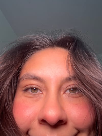 The image is a close-up, low-angle shot of a person's face, focusing on their eyes, nose, and cheeks, framed by dark hair. The lighting appears to be artificial, casting a warm glow on the skin and creating subtle shadows. The background is a dark, out-of-focus gray surface, possibly a wall or ceiling. There is no discernible information in the image that would allow for the determination of its location within San Luis, Peru, or any other city.