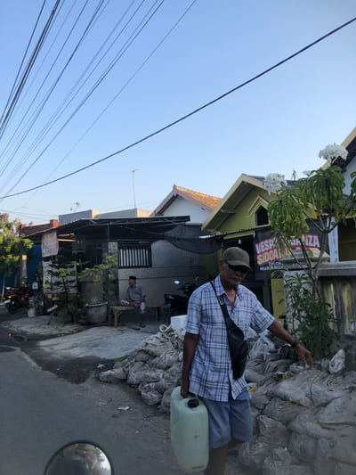In Kalidawir, Indonesia, a man wearing a plaid shirt, shorts, a baseball cap, and sunglasses walks on a paved street. He carries a large, pale green plastic container with a black lid in his right hand. A black sling bag is worn across his chest. To his right is a large pile of grey, lumpy sacks, possibly sandbags. Behind the man are houses with colorful facades and tiled roofs, some with awnings. A tree with white flowers is visible in front of one of the houses, adorned with a black banner that reads "VESPA SIDOARJO EXPLORERS NGABURIDE". A second man, wearing a grey shirt and checkered shorts, sits on a wooden bench in front of a house on the left. The sky is clear and blue, with numerous power lines crisscrossing overhead. Various motorcycles and potted plants are scattered throughout the scene. The overall impression is of a residential street scene in a tropical climate.