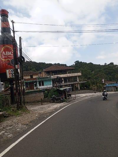 This image depicts a street scene in Ciloto, Indonesia, under an overcast sky. In the foreground, an asphalt road with a white solid line on its left edge curves gently to the right. On the right side of the road, a single individual wearing a helmet and a light-colored jacket is operating a white scooter, traveling away from the viewer.
To the left of the road, on an unpaved shoulder, two motorcycles are parked. Adjacent to these motorcycles are small, simple structures with green roofs, possibly roadside vendors or shelters. Behind these structures, a complex of multi-story buildings is visible, set against a backdrop of dense green foliage. One prominent building features a reddish-brown tiled roof and multiple levels with open-air balconies, appearing weathered. A lighter blue building is situated lower and to its left.
Dominating the left side of the frame is a colossal replica of a red and black "ABC Kecap Manis" (sweet soy sauce) bottle, functioning as an advertisement. This bottle is affixed to a utility pole structure from which numerous overhead power or communication lines extend horizontally across the upper portion of the scene. The distant background is characterized by lush green hills covered in trees.