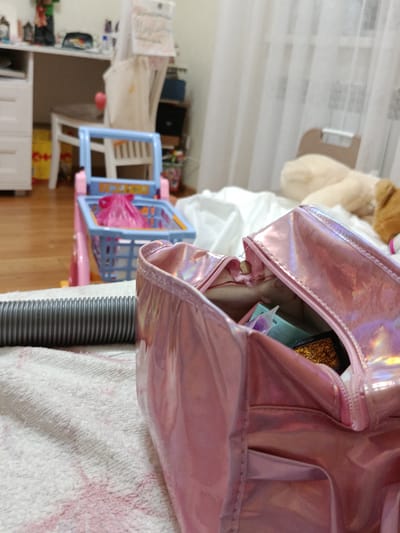 The image captures a close-up view of a pink holographic cosmetic bag with an open zipper, revealing some contents like eyeshadow. To the left, a gray, corrugated hose is visible, lying on a textured white and pink cloth. In the background, a child's toy shopping cart with a blue basket and pink frame is positioned. Further back, a white chair and a desk with drawers are seen, along with a white curtain. The scene appears to be indoors, within a domestic setting. The location of Novotitarovskaya, Russia, is not discernible from the visual information provided in the image.