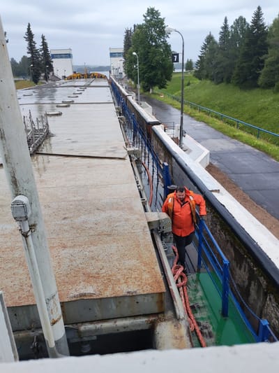 A man wearing an orange life vest and black pants walks along a walkway next to a canal lock. The walkway has a blue railing. A thick, red rope is coiled on the walkway. In the background, there are trees and buildings with white walls and blue trim. The sky is overcast. The location is the Белоусово canal lock, located in Белоусово, Russia.