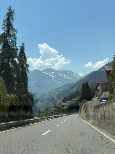 The image depicts a scenic mountain road in Troistorrents, Switzerland. A paved road with white lane markings curves to the right, bordered by a metal guardrail on the left. Beyond the guardrail, a steep, forested slope descends into a valley dotted with buildings. The valley floor appears to be a small village or settlement.

In the background, majestic snow-capped mountains rise against a clear blue sky with fluffy white clouds. The sunlight suggests it is daytime, likely mid-morning or afternoon. The overall atmosphere is one of natural beauty and tranquility, with the road suggesting travel and the possibility of exploration. No people are visible, but a dark-colored car is parked on the side of the road near some buildings higher up the slope. The architecture of the buildings appears to be traditional Swiss style, with dark roofs and wooden elements. The presence of the road and the guardrail indicates this is a well-maintained route, likely a popular tourist destination or a local thoroughfare through the mountainous region.