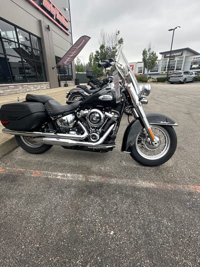 The image shows a black Harley-Davidson Heritage Classic motorcycle parked on asphalt outside a dealership in Township of Langley, Canada. The dealership building, with "Harley-Davidson" branding, is visible in the background. Also in the background is a "Holeshot" dealership and a silver SUV. The sky is overcast. The motorcycle is the primary subject, featuring chrome accents, saddlebags, and a windshield. An orange line is painted on the asphalt in the foreground.