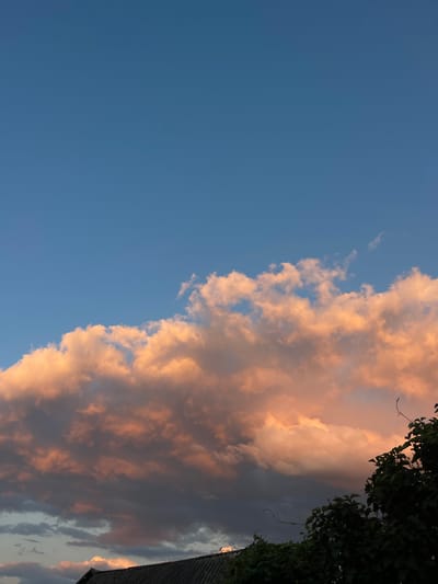 The image depicts a twilight sky over Krasnokutsk, Ukraine. A large, fluffy cloud formation dominates the middle ground, tinged with warm, peachy hues from the setting sun. Below the clouds, the silhouette of a dark, textured roofline is visible, likely belonging to a residential building. To the right, the dark green foliage of trees or bushes encroaches on the frame, their leaves catching faint light. The sky above is a clear, deep blue, transitioning from lighter shades near the horizon to a richer tone higher up. No people or animals are present in the scene.