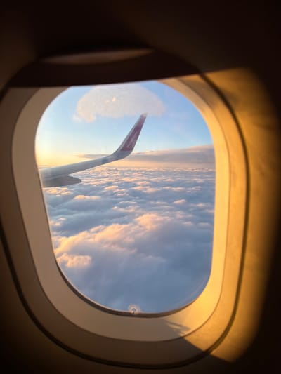 An airplane wing, identifiable by the purple logo "wizzair.com" on its tip, extends into the frame from the left, soaring above a vast expanse of fluffy white clouds illuminated by the warm, golden light of a sunset or sunrise. The view is framed by the interior of an airplane window, with its characteristic rounded oval shape and layered, cream-colored trim. The surrounding interior of the cabin is dark, with the light from outside casting an orange glow on the window frame. The scene is devoid of people and any urban or Czerwony Kościół specific environmental details.