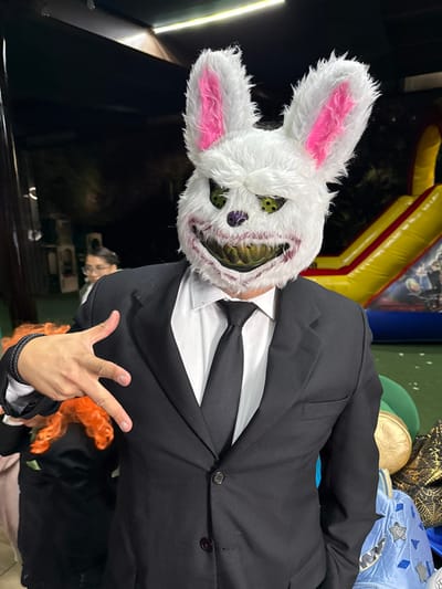 A person wearing a white, menacing rabbit mask with pink ears, yellow eyes, and sharp teeth. They are wearing a black suit with a white shirt and a black tie. The individual is making a hand gesture, and there is an orange, feathery object near their hand. The background features a dark setting with an inflatable structure in the distance.