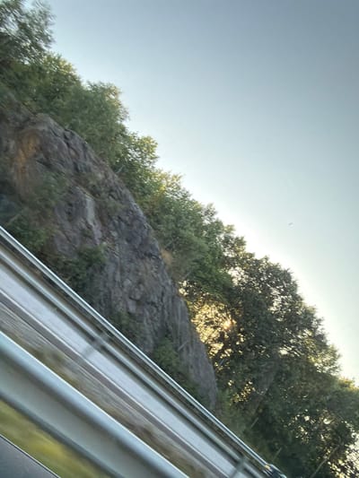 The image is an angled, close-up, outdoor shot of a rocky cliff face covered in green foliage. The sky is a pale blue and takes up the top portion of the frame. A metal guardrail runs across the bottom of the image, obscuring the foreground. The overall impression is that the photo was taken from a moving vehicle. The image does not contain any identifiable landmarks or features that would allow for the specific location within Kode, Sweden to be determined. There are no people or animals visible in the image.