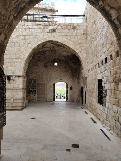 The image depicts an interior passage or courtyard within the Antipatris Fortress (Tel Afek National Park) in Rosh HaAyin, Israel. The structure is predominantly constructed from light-colored, rough-hewn stone blocks.

Two large, curved stone arches frame the immediate foreground, opening into a paved area floored with light-colored flagstones. On the far left, a stone pillar supports a circular mounted fixture, and a gray cylindrical waste bin is situated nearby. A metal informational screen with text and decorative patterns is partially visible on the extreme left. Further into the passage, a decorative, latticed wooden screen or door stands against the left wall.

A prominent, pointed stone archway centrally dominates the midground, serving as an exit to an exterior environment. Above this arch, a small rectangular opening is visible. Through this archway, approximately 5-7 individuals, dressed in casual, light-colored clothing, are seen standing or walking on a distant bright green grassy hill under a blue sky with scattered clouds.

The right wall of the passage is also built from light stone blocks and features a horizontal row of approximately ten small, rectangular apertures at an elevated height. Below these, two modern-style windows protected by black metal grates are installed. Dark, linear drainage channels are embedded in the flagstone floor along this wall. In the central area of the flagstone floor, several dark, square or rectangular metal plates are visible. Above the central arch and extending along the upper sections of the surrounding walls, an additional level of stone structure is apparent, topped with a black metal safety railing.