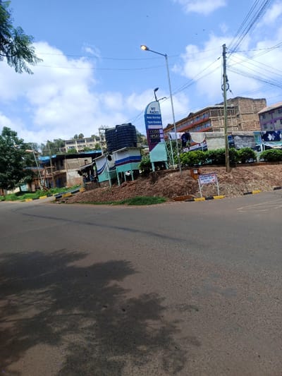 This image depicts a daytime scene in Murang'a Town, Kenya, specifically an area near the MT BACCO Auto Service Tyre Centre. The foreground features an asphalt road, partially shaded by unseen trees on the left, curving to the right. Yellow and black painted kerbstones line the right edge of the road, bordering a reddish-brown dirt embankment.

On the embankment, a vertical, multi-colored sign for "MT BACCO Auto Service Tyre Centre" is prominent, displaying "Auto Service" and "Tyre Centre" with numerical values "132.00" and "185.00." Behind the sign, two large black water tanks are mounted on a raised structure adjacent to a light blue and white service building. To the right of the sign, a street light pole with an illuminated lamp stands, and further right, a wooden utility pole supports multiple overhead power lines. A white sign with black text, "MERCANDISING AND BUSINESS ONLY ALLOWED," is affixed to the lower part of the embankment.

The midground and background consist of a densely built-up area on sloping terrain, featuring numerous multi-story buildings of varying construction styles, including some with exposed brick or concrete and others with painted facades, such as one prominent building with blue balconies. Green foliage is visible on the left side, complementing the partly cloudy blue sky overhead. No individuals or specific actions are distinctly identifiable within the scene.