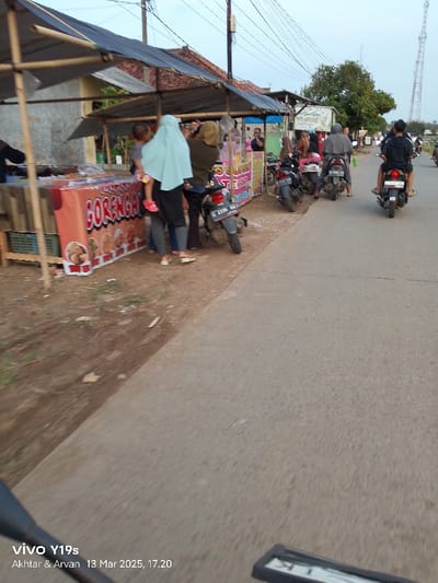 The image shows a street scene in Kecamatan Gabuswetan, Indonesia. It appears to be a street vendor market with several stalls set up along the side of the road. The stalls are covered with tarpaulin canopies and are selling various goods, including food items. There are several people in the scene, some are customers browsing the stalls, and others are vendors. There are also several motorcycles parked along the side of the road. The scene is bustling with activity, and the weather appears to be clear and sunny. The image is taken from the perspective of a person riding a motorcycle. The image appears to have been taken on March 13, 2023, at 5:20 PM.