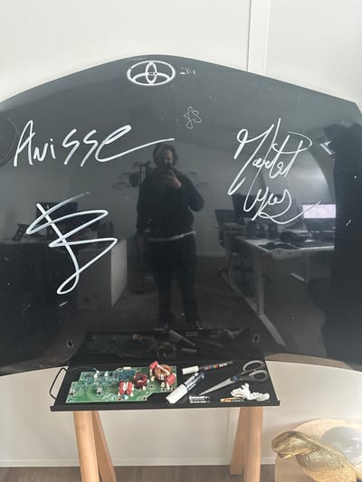 The image depicts an indoor scene in Thiais, France, focused on a detached, glossy black car hood positioned vertically against a white wall. The hood features a silver Toyota emblem centered at the top and is covered with white marker signatures. On the left side, "Anisse" is written above a large, stylized 'B' or '3'. On the right side, "Matteo Yos" is inscribed, with a faint "fs" above it.
A person, identified by their reflection in the hood, is visible wearing a dark hooded sweatshirt, dark pants, and a white undershirt. This individual, who has dark, wavy hair and a beard, is holding a smartphone, capturing the image. The reflection also reveals an interior office or workshop environment containing multiple desks with computer monitors and office chairs against white walls.
In the foreground, a black rectangular tray rests on a light-colored wooden A-frame stand. The tray holds a green circuit board with numerous electronic components, including inductors and capacitors. Also on the tray are two white marker pens (one appearing to be Posca brand), a pair of silver scissors with black handles, several small cylindrical items (possibly batteries or empty marker cases), and a crumpled piece of white material. To the lower right, a golden, stylized bird sculpture and a clear spherical object are visible on a light-colored surface. A light wood floor is visible at the bottom of the frame.