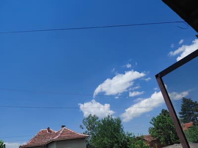 The image depicts a clear blue sky with scattered white clouds, observed from a low angle. In the foreground, a wire with a circular loop in the middle stretches across the upper portion of the frame. Below, the tiled roofs of several houses and dense green trees are visible. To the right, a dark-framed screen, possibly part of a window or door, is positioned at an angle, reflecting the sky and some of the surrounding environment. The scene appears to be in a rural or semi-rural setting, consistent with the village of Malorad, Bulgaria. There are no people or animals visible in the image.