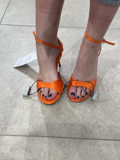 The image shows a view of two feet wearing orange open-toe high-heeled shoes with security tags still attached. The toenails are painted black. One ankle has a tattoo. The person is standing on a light gray tile floor. A tag or receipt is lying near one shoe. The location is Саларьево, Russia.