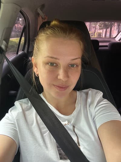 A young woman with blonde hair tied in a bun is sitting in the driver's seat of a car. She is wearing a white t-shirt and a black seatbelt is across her chest. Her left earring is visible. The background shows buildings and trees outside the car windows. The location is identified as Zelonaya Roshcha, Russia.