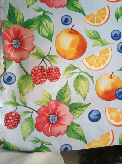 This is a close-up, overhead shot of a fabric with a vibrant, repeating pattern of fruits and flowers. The pattern features red flowers with blue centers, bright orange apples, orange slices, red raspberries, and blue blueberries, all interspersed with green leaves. The fabric is light blue. The image is cropped to show the texture and detail of the fabric, with no visible background elements that would indicate a specific location within Istaravshan, Tajikistan.
