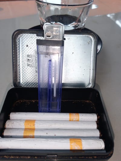 A close-up, top-down view of a black metal cigarette case containing several cigarettes is displayed on a light-colored, possibly concrete, surface. The case is open, revealing the cigarettes arranged horizontally. A clear plastic lighter with a blue interior is positioned vertically in the center of the open case, resting on top of the cigarettes. Behind the lighter, the silver lid of the cigarette case is visible, decorated with a patterned texture. A clear glass, partially filled with a dark liquid, sits on top of the lid. The overall environment appears to be outdoors or in a well-lit indoor space. The image does not provide specific location details within Kruengmanyang, Indonesia.