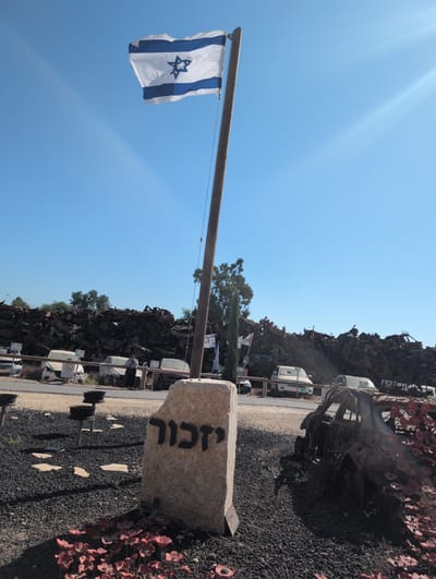 In the city of Sdot Negev Regional Council, Israel, an Israeli flag flutters atop a wooden pole, set against a clear blue sky. In the foreground, a stone monument engraved with the Hebrew word "Yizkor" (meaning "remember") stands amidst a bed of red artificial poppies. To the right, a burnt-out car, also adorned with red poppies, is partially visible. In the background, a vast expanse of discarded and rusted vehicles is piled high, forming a stark landscape under the bright sun. The scene appears to be an outdoor memorial or a junkyard with a solemn commemorative element.