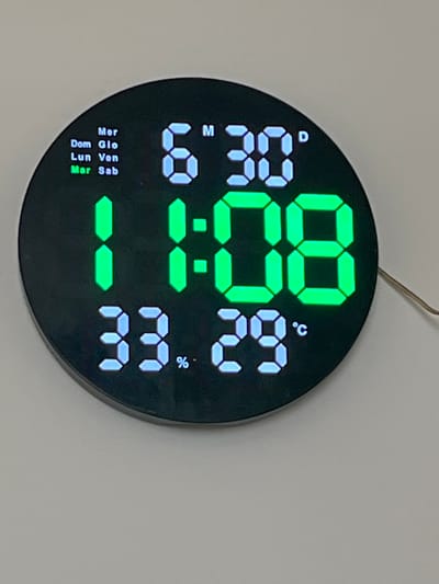 This is a close-up, eye-level shot of a digital clock mounted on a light grey wall. The clock is circular and black, with a green digital display showing the time as 11:08. To the left of the time, the clock displays "6:30" in blue digital numbers and the letter "M". Above the "6:30" is a small blue "D". To the right of the time, the clock displays "33.29" in blue digital numbers, indicating a temperature of 33.29 degrees Celsius, as denoted by the "°C" symbol. There is also a "%" symbol near the temperature display.
Along the bottom left edge of the clock, a series of days of the week are listed in Italian in blue digital text: "Dom", "Lun", "Mar", "Mer", "Glo", "Ven", "Sab". The "Dom" light is illuminated, indicating that it is Sunday. A thin, silver wire extends from the top center of the clock to the upper right. The overall scene is simple and focuses on the functional details of the digital clock. No people are visible, and the setting is a plain wall, suggesting an indoor environment.