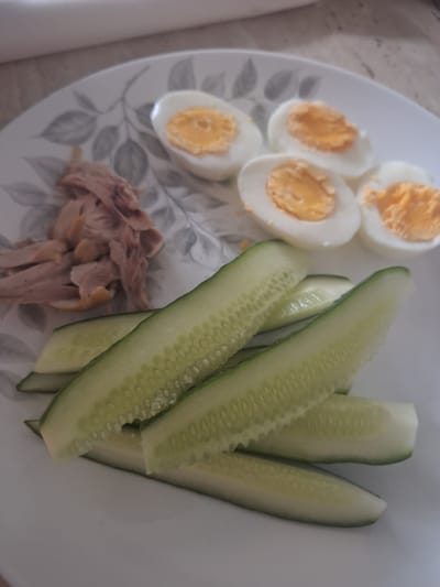A white ceramic plate with a grey floral pattern holds a meal consisting of sliced hard-boiled eggs, shredded chicken, and cucumber slices. Four halves of hard-boiled eggs, with their yellow yolks and white whites, are arranged in the upper right portion of the plate. To their left, a pile of shredded, light brown chicken is present. The lower half of the plate is covered with numerous lengthwise slices of cucumber, showcasing their green skin and white flesh with visible seeds. The plate rests on a light-colored, possibly marble or laminate surface. The lighting suggests an indoor environment, potentially a dining area. There are no people visible in the image, and no specific details indicate the location within Mińsk Mazowiecki, Poland.