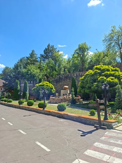 The image depicts a scene outside a wine cellar in Milestii Mici, Moldova. In the foreground, a cracked asphalt road with white and red pedestrian crossing markings is visible. To the right, a wooden signpost with several directional arrows points towards "Wine Tour" and "Info Center". Behind the road, a manicured lawn is bordered by a low, brick-faced wall topped with terracotta. Beyond this, a garden area features neatly trimmed boxwood bushes, ornamental trees, and lampposts. In the background, a stone wall, characteristic of a fortification, rises behind the garden. A wooden barrel is perched atop a stone structure, and wine glasses are displayed on another stone display. The overall environment is lush with various trees, including cypress and deciduous varieties, under a clear blue sky with scattered white clouds. No people are present in the image.