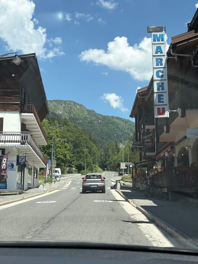 The image captures a sunlit street in Vallorcine, France, on a clear day with fluffy white clouds against a bright blue sky. The street is paved, with white lane markings and speed limit signs indicating "30". On the left side of the street, a multi-story building features a wooden facade on its upper levels and white painted lower levels with balconies. Shops are visible on the ground floor, with signage for "ON Running" and "LOCATION RENT". On the right side, a vertical sign with blue lettering spells out "MARCHE" ("Market") with "OUVERT" ("Open") at the top. Further down the street, more buildings with wooden exteriors house businesses including a "Stone BAR PIZZERIA" and a "L'Étranger" establishment. A car is driving away from the camera down the street, and on the right sidewalk, bicycles are parked outside a wooden establishment. Lush green trees and a forested mountain slope form the backdrop to the scene.