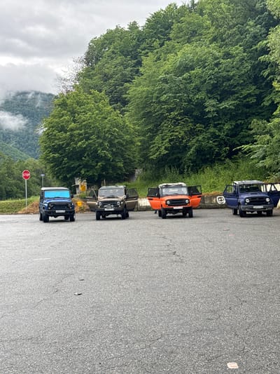 Four off-road vehicles are parked on an asphalt surface in front of a dense forest. From left to right, the vehicles are a blue Jeep-like SUV, a tan SUV with its doors open, an orange SUV with its doors open, and a dark blue SUV with its doors open. A stop sign is visible in the background to the left, and mountains covered in mist and trees are visible in the distance. The sky is overcast. The scene appears to be in a natural or semi-natural environment, possibly on the outskirts of a settlement.