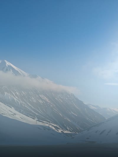 A sweeping panoramic view captures a majestic mountain landscape, predominantly covered in snow, under a clear blue sky. The scene is dominated by massive, rugged peaks, their upper reaches stark white with snow, contrasting with the darker rock faces and valleys below. A band of wispy white clouds or mist partially obscures the mid-section of the mountains, adding a sense of depth and ethereal beauty.
The sky is a vibrant, unbroken blue, occupying the upper half of the image, suggesting bright daytime conditions. Towards the upper right, a soft glare indicates the presence of the sun, though it is not directly visible. Long shadows are cast across the lower mountain slopes and valleys, plunging them into a deep, cool grey, while the upper peaks are bathed in light.
In the lower-left quadrant, two electricity pylons (power line towers) stand on a snow-covered slope, providing a subtle hint of human presence amidst the otherwise untouched natural grandeur. The overall impression is one of vastness, tranquility, and the raw, cold beauty of a high-altitude, snowy environment. No visible text is present. Given the context, this scene likely depicts a mountainous region near Ganisi, Georgia.