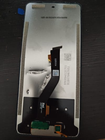 A damaged smartphone screen is lying on a dark wooden surface. The screen is detached from the phone and displays a silvery backing with some black adhesive strips. Several labels and codes are visible on the screen's surface. A blue object, possibly a piece of plastic, is located in the upper left corner of the image. The overall condition of the screen suggests it has been removed from a device and may be in the process of repair or replacement. There is no information within the image to indicate a specific location in Mělník, Czechia.