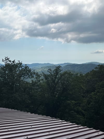 This media file captures a scenic view of rolling, tree-covered hills under a cloudy sky. The foreground features a section of a corrugated metal structure, likely a roof or wall, with distinct vertical ridges. To the left of the structure, a dense canopy of dark green trees is visible, their leaves creating a rich texture. Beyond the immediate trees, layers of distant hills recede into a hazy, blue-green landscape, suggesting a considerable distance and atmospheric depth. The sky is filled with a mix of dramatic, dark gray clouds and patches of brighter, lighter gray and white clouds, with some hints of blue sky peeking through. The sun appears to be casting a bright light, possibly from the upper left, illuminating some of the clouds and casting a soft light on the scene. The overall impression is one of a natural, slightly moody landscape, possibly captured in the late afternoon or early evening due to the soft lighting and cloud cover. The location is given as Voronstovka, Russia, which places this scene within a geographically diverse region of the country. There is no visible text in the image.