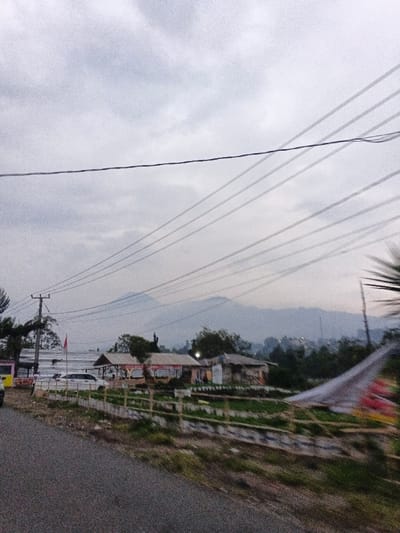 The scene depicts a rural or semi-rural landscape under an overcast sky, presented as being located in Phước Hòa, Vietnam. A grey asphalt road occupies the lower left foreground. A yellow vehicle is partially visible on the far left edge of the road. In the mid-ground, a utility pole supports multiple overhead power lines that traverse the upper half of the image. Below the power lines, a series of modest buildings are visible, interspersed with vegetation. One building features a brown sloped roof, beneath which tables and chairs are faintly discernible, suggesting a dining establishment. A low, light-colored fence defines plots of cultivated ground in front of the buildings. A red and white horizontally striped flag is affixed to a pole near one of the structures on the left. In the background, hazy, blue-grey mountains rise against the cloudy horizon. No individuals or specific actions are discernible within the frame.