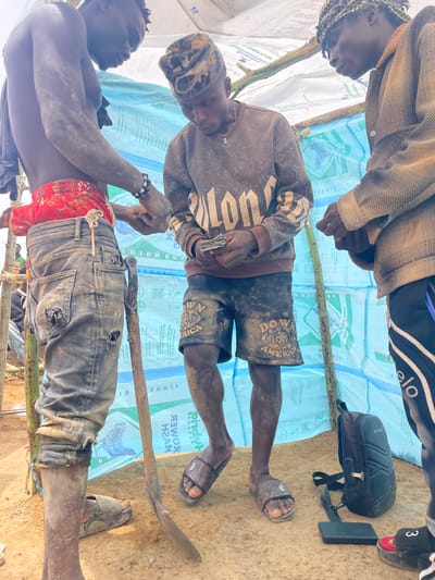 The image shows three partially visible dark-skinned men. One man, shirtless except for a red fabric and blue jeans with holes, appears to be working on something with a tool that has a blade and a handle. A second man is wearing a brown shirt with "DOW AMERICA" printed on it, and brown shorts, also with the words "DOW AMERICA" printed on them. This man is also holding a tool. The background is a makeshift shelter made of light blue plastic sacks with images of chickens and text printed on them. Parts of the sacks are visible, including the words "HYBRID", "KOMEK", "HABKID", "DYNOCOL," and "AUO MUIME". A bag and flip-flops are on the ground, suggesting they are inside a shelter. The overall environment appears dusty and possibly rural. Location is Maiyanga, Nigeria.
