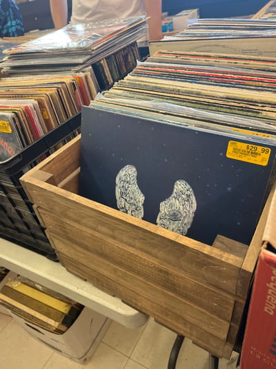The image displays an indoor scene within Hometown Pickers, located at 1200 S Broadway, Edmond, Oklahoma, United States. The primary focus is on a collection of vinyl records. In the foreground, a prominent dark blue vinyl record album with abstract white winged figures on its cover is positioned upright within a brown wooden crate. A yellow price sticker on this album indicates "USED $29.99" and "HOMETOWN PICKERS". To the immediate left, a black plastic crate is filled with numerous other vinyl records, some displaying similar yellow price tags. Stacks of additional vinyl records, many enclosed in clear plastic sleeves, are visible on a white folding table extending into the background. Partially visible in the background are two individuals; one wearing a white t-shirt stands behind the table, and another wearing a blue patterned shirt is further to the left. A red and white cardboard box is partially visible on the right side of the frame, and a white plastic bin is visible under the table on the left. The flooring is a light-colored tile, and the area is brightly illuminated.