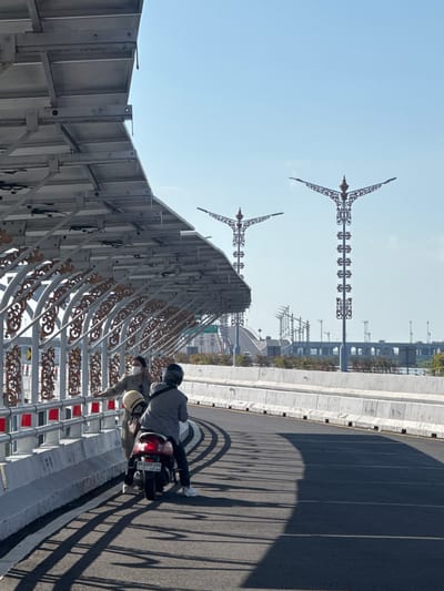 The image shows two people on a motorcycle, with one person standing beside them, on a bridge in South Denpasar, Indonesia. A covered walkway with ornate metal supports runs along the side of the bridge, casting long shadows on the asphalt road. Two decorative streetlights with intricate designs stand tall against a clear blue sky. In the background, other bridges and structures are visible, hinting at urban development. The motorcycle is red, and the license plate is visible. One person is wearing a plaid jacket, while the other is wearing a light-colored outfit and a face mask.