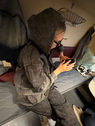 A person is lying on a bed, looking at their phone. They are wearing a hooded sweatshirt and jeans. The bed has a blue comforter and a patterned pillow. A suitcase is visible near the bed. The scene appears to be indoors, possibly in a train car. It is impossible to confirm the location of Tsibanobalka, Russia, from the visual data.