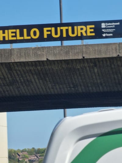 This outdoor image features a prominent, tall, vertical black banner displaying yellow text and logos against a clear blue sky. The main text, reading upwards, spells out "HELLO FUTURE" in large, uppercase letters. Above this, smaller white text and associated logos are visible, including "Gateshead Council" with its emblem (three geometric shapes), and below it, "Together for the North" alongside the logo for "bam".

To the right of the banner, a textured concrete structure, possibly a pillar or wall, shows a pattern of horizontal striations. Further to the right, a large, curved object in white and green is partially visible, which could be part of a modern building, vehicle, or sculpture.

In the background, under the clear blue sky and to the right of the concrete structure, a residential area is discernible, featuring several houses with brown roofs nestled amongst green trees. The overall setting suggests a bright, sunny day in an urban or semi-urban environment, identified as Gateshead, United Kingdom, by the banner's text. No people or specific activities are present in the scene.