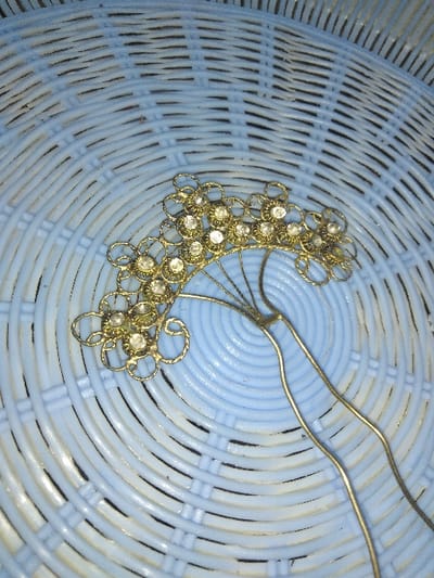 A gold-colored metal hair ornament with a U-shaped base rests on a light blue, concentric circular woven plastic surface, likely a basket or tray. The ornament features a cluster of interconnected floral or petal-like motifs, each containing a single clear, faceted stone or rhinestone. The design extends from a central point, forming an arc. No people, actions, or interactions are present in the scene. The image depicts an object in an indoor setting within Ottignies-Louvain-la-Neuve, Belgium.