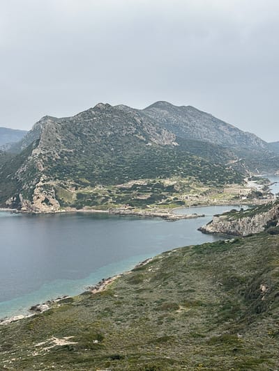 This panoramic image, taken in Datça, Turkey, captures a rugged coastal landscape under a hazy sky. The scene is dominated by a large, steep mountain range stretching across the left side of the frame, from the background towards the midground. Its slopes are densely covered with dark green vegetation, indicating a lush but rocky terrain.
A calm body of water, likely a bay or a strait, occupies the center and right side of the view. The water displays varying shades of blue and gray, with lighter, turquoise tones closer to the shoreline, suggesting shallow areas. Along the base of the left mountain range, a narrow strip of land extends into the water. This strip hosts a small settlement, characterized by a cluster of light-colored buildings and visible roads, possibly including a small port or dock area. Another heavily vegetated hillside, similar in appearance to the distant mountains but closer to the foreground, frames the right side of the image, sloping down towards the water's edge.
The sky is a soft, light gray, indicative of an overcast day or atmospheric haze, which diffuses the sunlight and creates a serene, somewhat muted light across the landscape. There are no people or specific activities visible, emphasizing the natural beauty and tranquility of the scene. No discernible text is present in the image. The time of day appears to be daytime, with soft lighting.