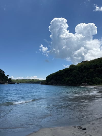 The image depicts a coastal scene in Sakti, Indonesia, showing a beach with sand to the right, turquoise water with gentle waves occupying the central and right portions of the frame, and a forested cliff rising along the shoreline. A large, puffy, white cloud formation stands out against a clear blue sky in the upper-left quadrant. The orientation of the image is vertical.