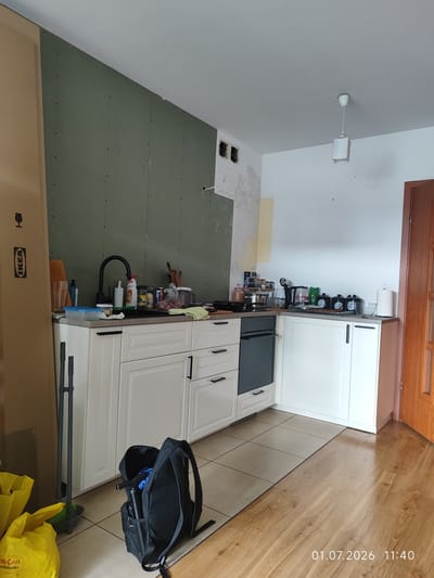 The image depicts an unfinished kitchen renovation. The walls show exposed drywall on the left, with a section covered in green drywall on the right. A white ceiling with a simple pendant light fixture is visible. The kitchen itself features white cabinets with black handles, a black sink and faucet, and a black oven. Various kitchen items, including pots, pans, bottles, and containers, are scattered on the countertops and sink area. A black backpack is open on the floor, containing what appears to be cleaning supplies. Yellow plastic bags and a broom with a dustpan are also present on the floor. A wooden door is visible on the right side of the frame. The floor is a mix of beige tiles and wooden planks. A timestamp "01.07.2026 11:40" is overlaid on the image. The scene is consistent with a residential kitchen undergoing renovation and is located in Lębork, Poland.