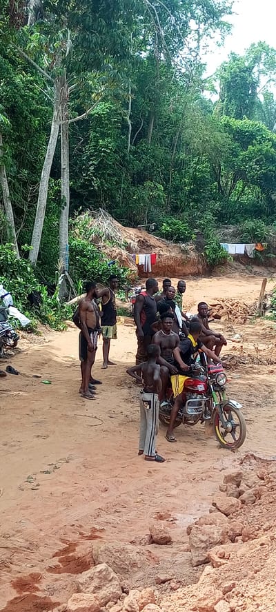 The image depicts a casual gathering of approximately 10-15 men and boys in a rural, forested setting in Aloko, Nigeria, during daylight hours. The ground is predominantly reddish-brown dirt, uneven and scattered with small rocks and debris.
In the midground, a man sits on a bright red motorcycle with distinct yellow accents on its wheels, wearing a black and yellow top and shorts. A young boy in grey sweatpants stands facing away from the camera, positioned in front of the motorcycle. Several other men, many shirtless or wearing tank tops and shorts, are standing and sitting around the area, engaging in what appears to be casual socialization or resting. Some individuals look towards the camera, with one man seated on the motorcycle displaying a relaxed smile.
The background features dense, vibrant green jungle vegetation and tall trees with light-colored trunks, indicating a heavily forested environment. A large mound of reddish-brown earth is visible in the mid-background, with some laundry hanging on a line or draped over it, suggesting a lived-in area. The lighting is bright and diffuse, consistent with an overcast or hazy day, and implies warm weather. No discernible text is visible within the image. The overall scene conveys a sense of a communal moment in a natural, unpaved outdoor environment.