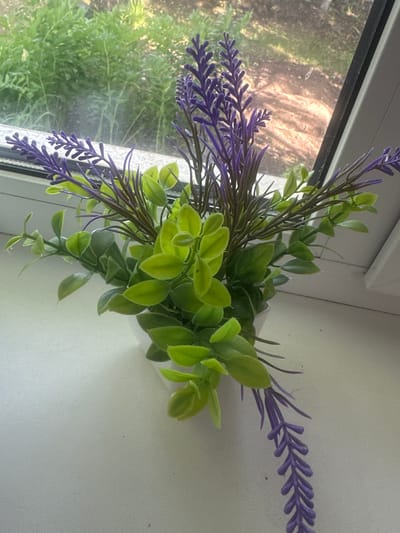 A close-up shot shows a faux bouquet of lavender and green foliage resting on a white windowsill. The artificial lavender stems are a vibrant purple with delicate, textured florets, interspersed with thin, dark green leaves. The green foliage consists of clusters of small, oval-shaped leaves in varying shades of light and medium green, with some stems drooping downwards.

Through the window on the left, a blurry background suggests an outdoor scene with lush green trees and vegetation, hinting at a daytime setting. The light filtering through the window casts a soft glow on the windowsill and the artificial plants, highlighting their texture and form. The overall impression is one of simple, decorative arrangement on a windowsill, possibly in a home or office. No people are visible in the image. No text is present.