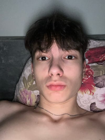 The image depicts a young individual, likely male, lying supine indoors, possibly in a bed, in the city of Zainsk, Russia. The subject, possessing fair skin, dark brown hair with a fringe, and dark brown eyes, gazes directly into the camera. A silver-colored chain necklace with a small pendant is worn around the neck. The individual's upper chest and collarbones are visible, indicating a shirtless state. The head rests on a pillow featuring a floral pattern with red, pink, purple, and green elements against a lighter background. A plain, dark grey or dark green surface is visible to the left behind the subject's head, suggesting a wall or headboard. The lighting is dim, casting soft shadows. No other individuals or significant objects are discernible.