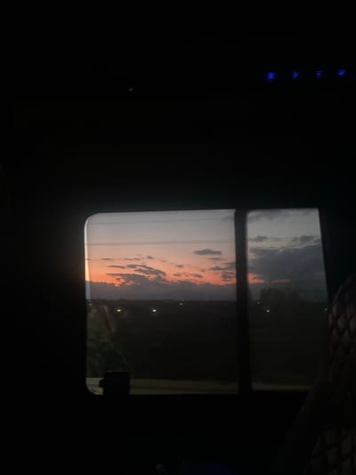 This image captures a dusk scene viewed through a vehicle's window, likely in the city of Akhalsheni, Georgia. The primary focus is the sky, painted with hues of orange and pink from the setting sun, with scattered dark clouds adding texture. Below the sky, a dark landscape is visible, dotted with faint lights that suggest distant buildings or streetlights. The window frame is a prominent feature, dividing the view into two panes. The dark interior of the vehicle is also visible, with a hint of a patterned seat on the right side. The overall impression is one of quiet observation during twilight.
