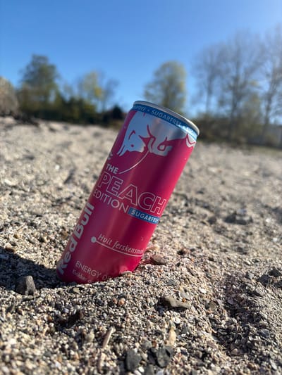 The image, oriented diagonally, presents a close-up view of a Red Bull "The Peach Edition" can resting on a textured ground composed of small, varied-colored pebbles and crushed shells. The can is predominantly a vibrant pink/red, featuring white and blue text, and a silver top. It appears to be slightly settled into the gravelly surface.
In the background, a bright, clear blue sky occupies the upper portion of the frame, indicating a sunny day. Out-of-focus, silhouetted trees with light or bare branches are visible against the sky, suggesting an outdoor, open environment.
No activity or people are depicted; the scene is static. The strong, natural light illuminates the can and the ground, highlighting the gravel's texture. The setting is likely an outdoor area, possibly a beach or shoreline in Nærsnes, Norway, given the appearance of the ground and the open sky.
Visible text on the can includes "SUGARFREE" (repeated), "THE PEACH EDITION," "Red Bull," "Hvit ferskensmak" (Norwegian for "White peach flavor"), "ENERGY DR" (part of "ENERGY DRINK"), and "Kvikker op" (Norwegian for "Refreshes" or "Enlivens").